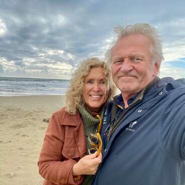 Una pareja de personas mayores sonriente posa muy unida en una playa de arena, con un cielo nublado sobre ellos y las olas del mar a sus espaldas. Parecen felices, disfrutando de un día fresco y pintoresco al aire libre.