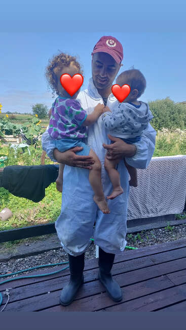 Un homme vêtu d'une combinaison bleu clair et d'une casquette rouge se tient debout à l'extérieur, tenant tendrement deux jeunes enfants dont les visages sont recouverts d'émojis en forme de cœur, entourés de verdure.