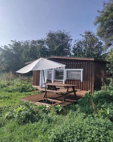 Une petite cabane en bois se dresse au milieu d'un pré verdoyant. Une table de pique-nique et un grand parasol invitent à la détente sur la terrasse ensoleillée et paisible à l'extérieur.