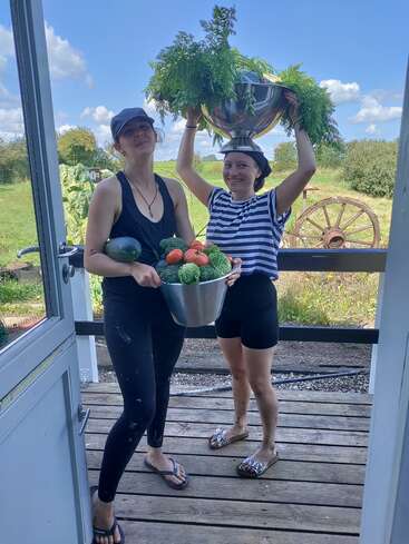 Deux femmes se tiennent sur un porche en bois, souriantes, tenant des bols remplis de légumes fraîchement récoltés. L'une d'elles porte un bol en équilibre sur la tête. Elles sont entourées d'un paysage rural.