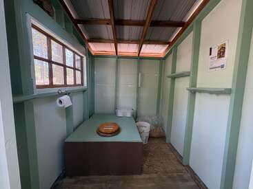 This image shows a rustic, open-roof composting toilet with a wooden seat, toilet paper, window, light green walls, and buckets of sawdust inside.