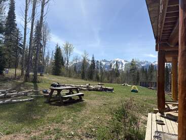 A rustic cabin porch overlooks a grassy yard with picnic table, logs, scattered belongings, and trees. Snow-capped mountains rise majestically under a bright blue sky.