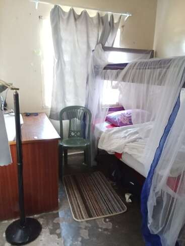 Ein kleines, einfaches Zimmer mit einem Etagenbett mit Moskitonetz, einem Plastikstuhl, einem Schreibtisch, einer Lampe, einem gestreiften Teppich und hellen Vorhängen am Fenster.