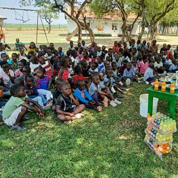 Eine große Gruppe Kinder sitzt ganz aufmerksam auf dem Rasen unter Bäumen und schaut auf einen Tisch mit Getränken. Im Hintergrund sieht man einen Spielplatz und ein Gebäude.