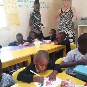 De jeunes enfants sont assis à des tables colorées, concentrés sur leurs dessins réalisés au marqueur. Deux enseignants les supervisent. Des collations sont disposées sur les tables et des citations inspirantes décorent les murs.