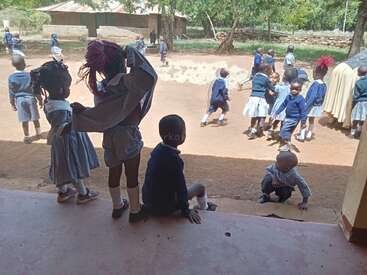 Ein paar kleine Kinder in Schuluniformen spielen draußen auf einem Sandplatz. Einige stehen am Eingang eines Gebäudes, während andere in Gruppen unter Bäumen rumtollen.