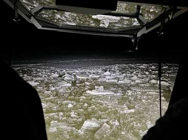 A imagem mostra um espaço escuro e fechado com vista para um corpo de água coberto de gelo e pedras, possivelmente tirada de um veículo ou abrigo.