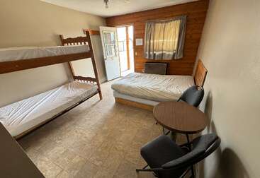 A imagem mostra um quarto com dois conjuntos de beliches, cada um com uma mesa e cadeiras, com uma parede branca e um piso cinza, provavelmente em um dormitório ou albergue.