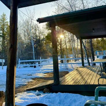 A imagem retrata uma cena serena de inverno com um deck de madeira com telhado, cercado por terra e árvores cobertas de neve, com o sol brilhando entre as árvores ao fundo.