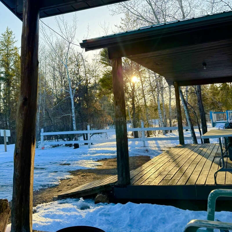 A imagem retrata uma cena serena de inverno com um deck de madeira com telhado, cercado por terra e árvores cobertas de neve, com o sol brilhando entre as árvores ao fundo.