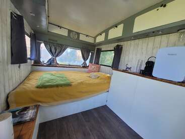 El interior de esta acogedora y compacta autocaravana cuenta con una cama grande con ropa de cama amarilla, cortinas en las ventanas, espacio de almacenamiento en la parte superior, superficies de madera, pequeños electrodomésticos y encantadores detalles decorativos por todas partes.