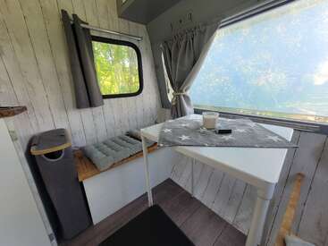El acogedor interior de esta autocaravana cuenta con una pared de madera blanca, un banco acolchado, una mesita con un mantel con motivos de estrellas, una vela y una gran ventana con cortinas grises.
