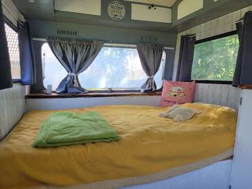 Una acogedora cama de caravana con una manta amarilla, una toalla verde, un peluche y una almohada con el letrero «Camping Queen». La luz del sol se cuela a través de las ventanas con cortinas, creando un ambiente tranquilo.