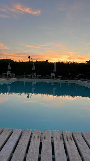 A imagem retrata uma cena serena de piscina ao pôr do sol, com um deck de madeira, espreguiçadeiras, guarda-sóis e uma cerca, em um céu vibrante com nuvens cor-de-rosa.