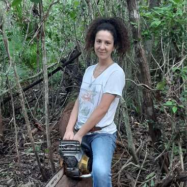 A woman with curly hair, wearing a white T-shirt and jeans, sits on a log in a dense forest, holding a chainsaw, smiling calmly.