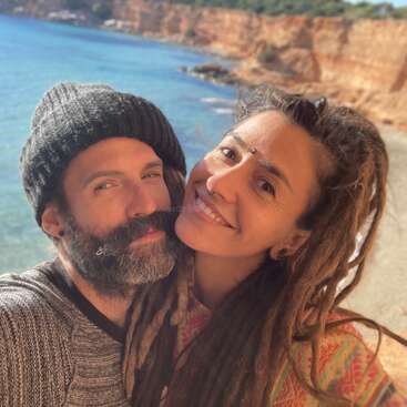 La imagen muestra a un hombre y una mujer posando para un selfie en una playa, con la cabeza de la mujer apoyada en el hombro del hombre, en un paisaje de acantilados.