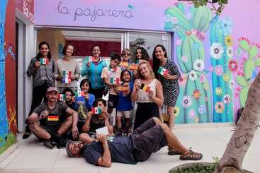 Eine bunte Gruppe von lächelnden Leuten und Kindern hält verschiedene Landesflaggen vor einem bunten Gebäude mit Kakteenwandmalereien und dem Schild „La Pajarera” darüber hoch.