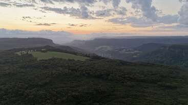 Un paysage à couper le souffle au coucher du soleil, avec des collines verdoyantes, des forêts denses, des montagnes lointaines et un ciel coloré avec des nuages épars, créant une atmosphère sereine et tranquille.