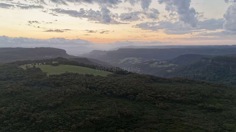 Un paysage à couper le souffle au coucher du soleil, avec des collines verdoyantes, des forêts denses, des montagnes lointaines et un ciel coloré avec des nuages épars, créant une atmosphère sereine et tranquille.