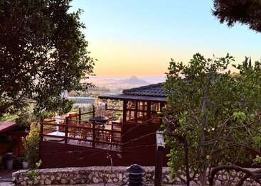 A cozy wooden house with a charming terrace sits among lush green trees, overlooking a scenic valley and mountains, illuminated by the warm, golden sunset.