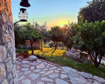 A beautiful stone patio leads to a lush garden with trees, a small fountain, and lantern. The sun sets warmly, creating a peaceful, serene atmosphere.