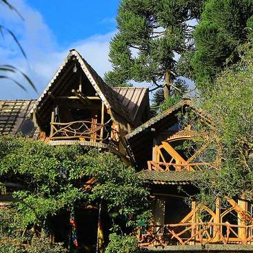The image depicts a charming wooden house with a sloping roof, nestled among lush greenery and trees, set against a serene blue sky with white clouds.