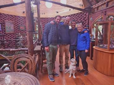 The image depicts four individuals, a dog, and a cat posing for a photo in a rustic room with wooden floors, walls, and ceiling, surrounded by various objects.