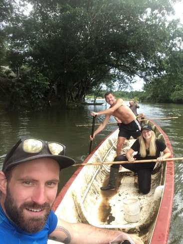 The image depicts a group of people in a canoe on a river, with one man taking a selfie in the foreground. The canoe is white with a red trim.