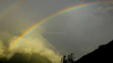 A vibrant rainbow-colored rainbow is prominently displayed in the sky, with its arches extending from the top of the image, set against a clear blue sky. The rainbow is surrounded by a light gray background.