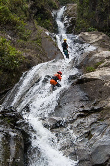Two people rappel down a rocky waterfall, wearing helmets and harnesses, with ropes attached to their waists, amidst lush greenery and large rocks.