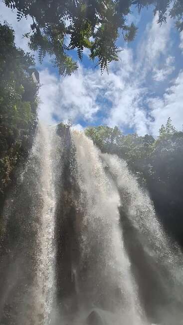 Powerful waterfall cascades down lush, rocky cliffs under a vibrant blue sky, framed by green foliage and mist. Sunlight highlights the dramatic, refreshing natural scene.