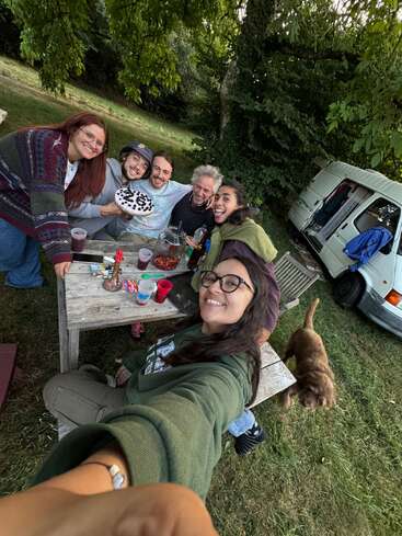 A group of six friends smile widely while gathered around a picnic table outdoors. There’s cake, snacks, drinks, a brown dog, and a white van in the background.