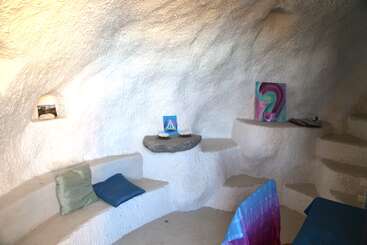 This cozy room has white, cave-like walls, built-in shelves, colorful pillows, abstract artwork, and a table with a blue cloth, creating a peaceful, artistic atmosphere.