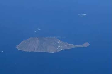 This aerial photo shows a small, rocky island surrounded by deep blue ocean water, with sparse vegetation and some white patches resembling snow or rocky outcrops.