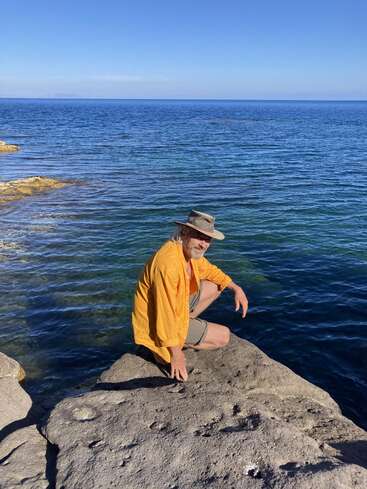 Une personne vêtue d'un t-shirt jaune et d'un chapeau est accroupie sur un rivage rocheux, au bord d'une mer bleue et calme, sous un ciel dégagé, profitant de la vue apaisante.