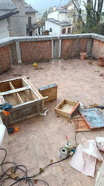 Une terrasse avec des outils éparpillés, des pots de peinture, des meubles en bois, des pots, un ballon, des feuilles mortes et des maisons de village en arrière-plan. La scène semble rustique.