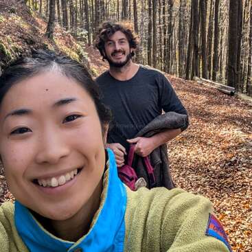 Une femme souriante prend un selfie avec un homme derrière elle sur un sentier forestier paisible et ensoleillé, couvert de feuilles d'automne. Tous deux ont l'air heureux en plein air.
