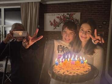 Two kids smiling and holding a birthday cake with lit candles spelling “HAPPY BIRTHDAY.” An adult takes a photo, everyone showing peace signs, cozy indoor setting.