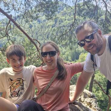 Three people smile together outdoors, surrounded by lush green trees and mountains. They appear happy, enjoying a sunny day and wearing casual clothes and sunglasses.