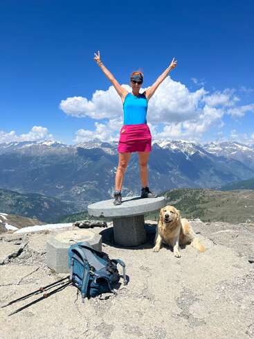 Une femme se tient debout, triomphante, sur une plate-forme de pierre, les bras levés en signe de victoire, avec son chien à ses côtés. Des montagnes majestueuses et un ciel bleu occupent l'arrière-plan.