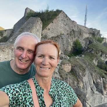 Un couple souriant prend un selfie devant une imposante forteresse de pierre érigée sur une colline rocheuse, au milieu d'un écrin de verdure sous un ciel légèrement nuageux.