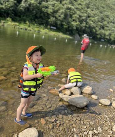 Zwei Kinder, die Schwimmwesten und Mützen tragen, spielen am Flussufer; das eine hält eine Wasserpistole in der Hand, das andere erkundet die Felsen. Etwas weiter entfernt steht ein Erwachsener.