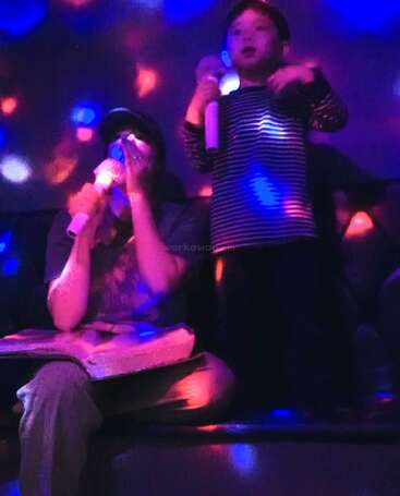 Ein Kind und ein Erwachsener singen gemeinsam Karaoke in einem schummrigen Raum mit bunten, leuchtenden Lichtern. Beide halten Mikrofone in der Hand, was für eine fröhliche, lebhafte Atmosphäre sorgt. Ein toller Abend.