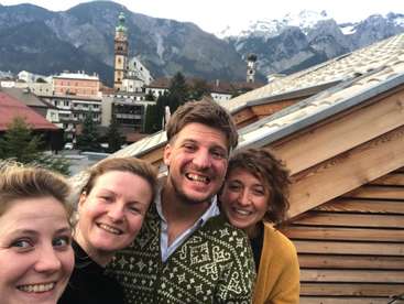 Das Bild zeigt vier Personen, die für ein Selfie auf einem Dach posieren, mit einer Stadt und Bergen im Hintergrund. Sie lächeln alle.