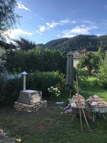 Das Bild zeigt eine ruhige Hinterhofszene mit einem kleinen Ofen im Freien, umgeben von üppigem Grün und einer malerischen Bergkulisse an einem sonnigen Tag.