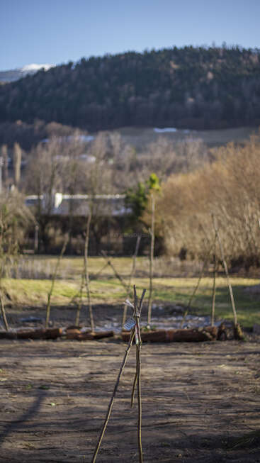 Das Bild zeigt eine rustikale Gartenszene mit einem Tipi im Vordergrund, umgeben von einer verschwommenen Landschaft mit Bäumen, Gras und entfernten Gebäuden.