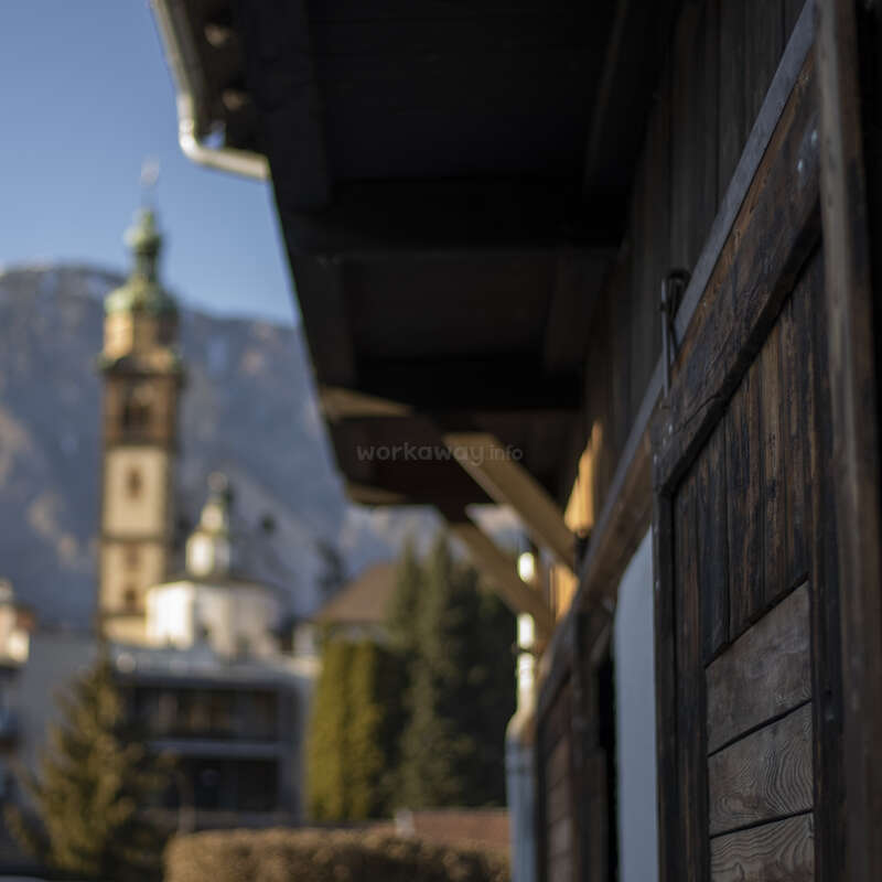 Das Bild zeigt ein hölzernes Gebäude mit schrägem Dach vor einem unscharfen Hintergrund mit einem Kirchturm und einer Berglandschaft unter einem klaren blauen Himmel.