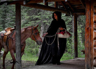 A woman in a black hooded cloak stands on a wooden porch, holding a basket, gently leading a saddled brown horse amidst a forest setting.
