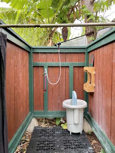 The image depicts an outdoor shower area with a wooden fence, a shower head, and a bucket, situated in a tropical setting with lush greenery surrounding it.