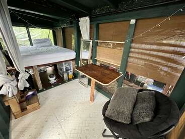 The image depicts a cozy room with a bed, desk, and chair, featuring a window with blinds and a mirror, surrounded by a serene natural setting.